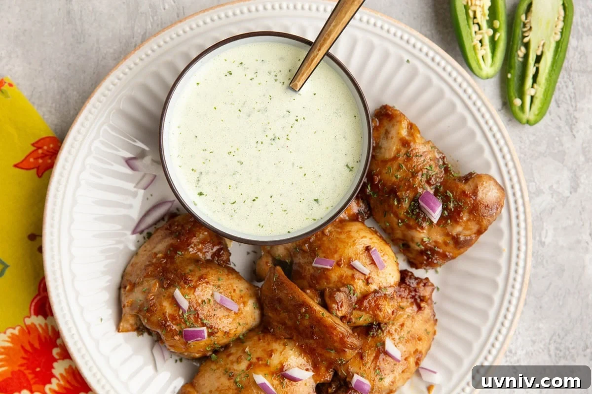 Peruvian Roast Chicken with Aji Verde Green Sauce Recipe 8 Peruvian chicken on a white plate with a bowl of green sauce and a sliced in half jalapeno to the side.