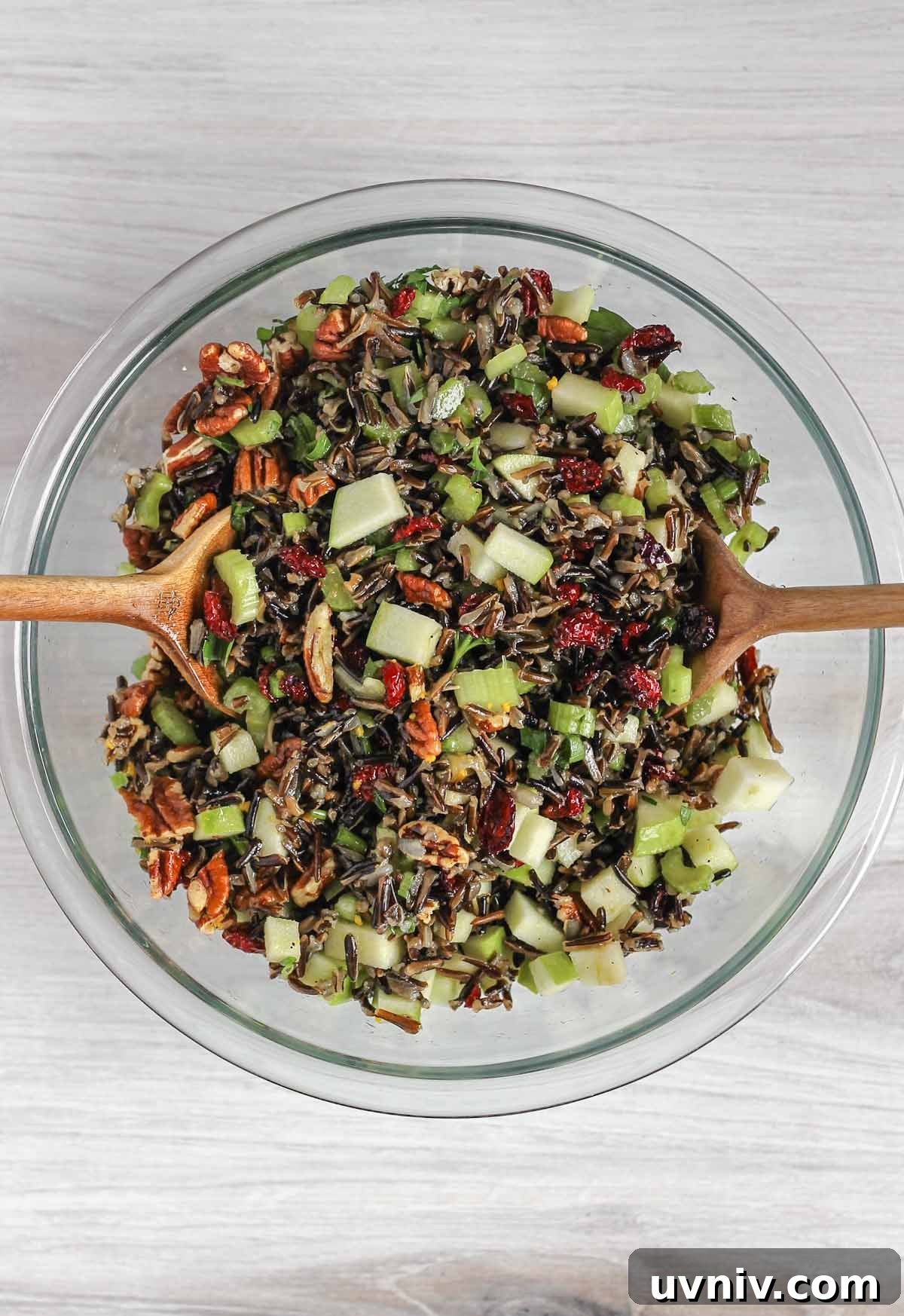 Wild Rice Salad with Cranberries & Pecans — Chilled Side Dish 9 Two wooden salad utensils mixing wild rice salad ingredients in a glass mixing bowl.