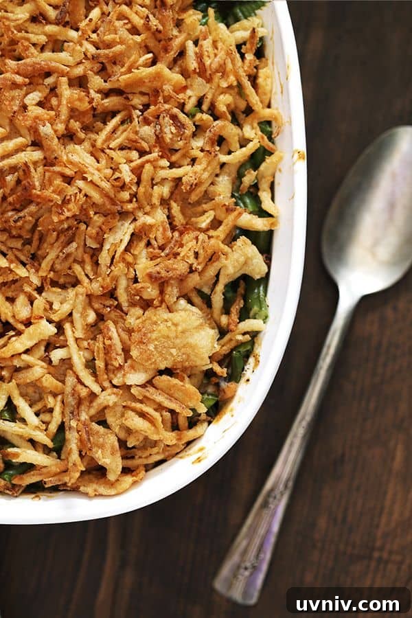 Twice the Onion Green Bean Casserole A white casserole dish of Twice the Onion Green Bean Casserole sits on a wooden surface with a spoon sitting to the right of it.