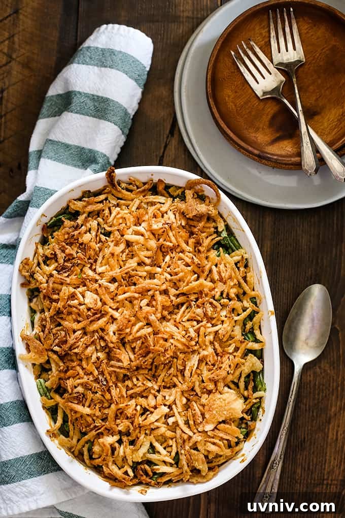 Twice the Onion Green Bean Casserole A white casserole dish of Twice the Onion Green Bean Casserole sits on a wooden surface surrounded by a striped napkin, spoon and a stack of plates.