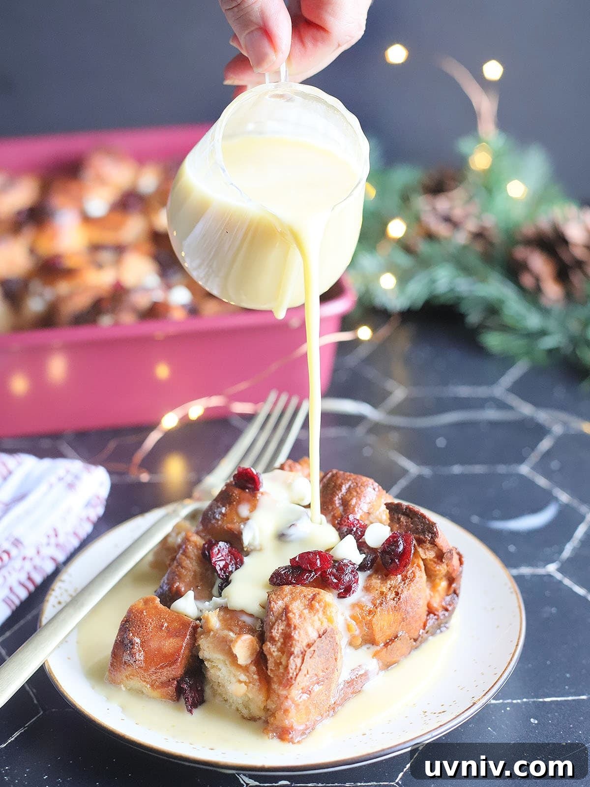 Pouring rum creme anglaise over the top of a slice of white chocolate cranberry bread pudding.