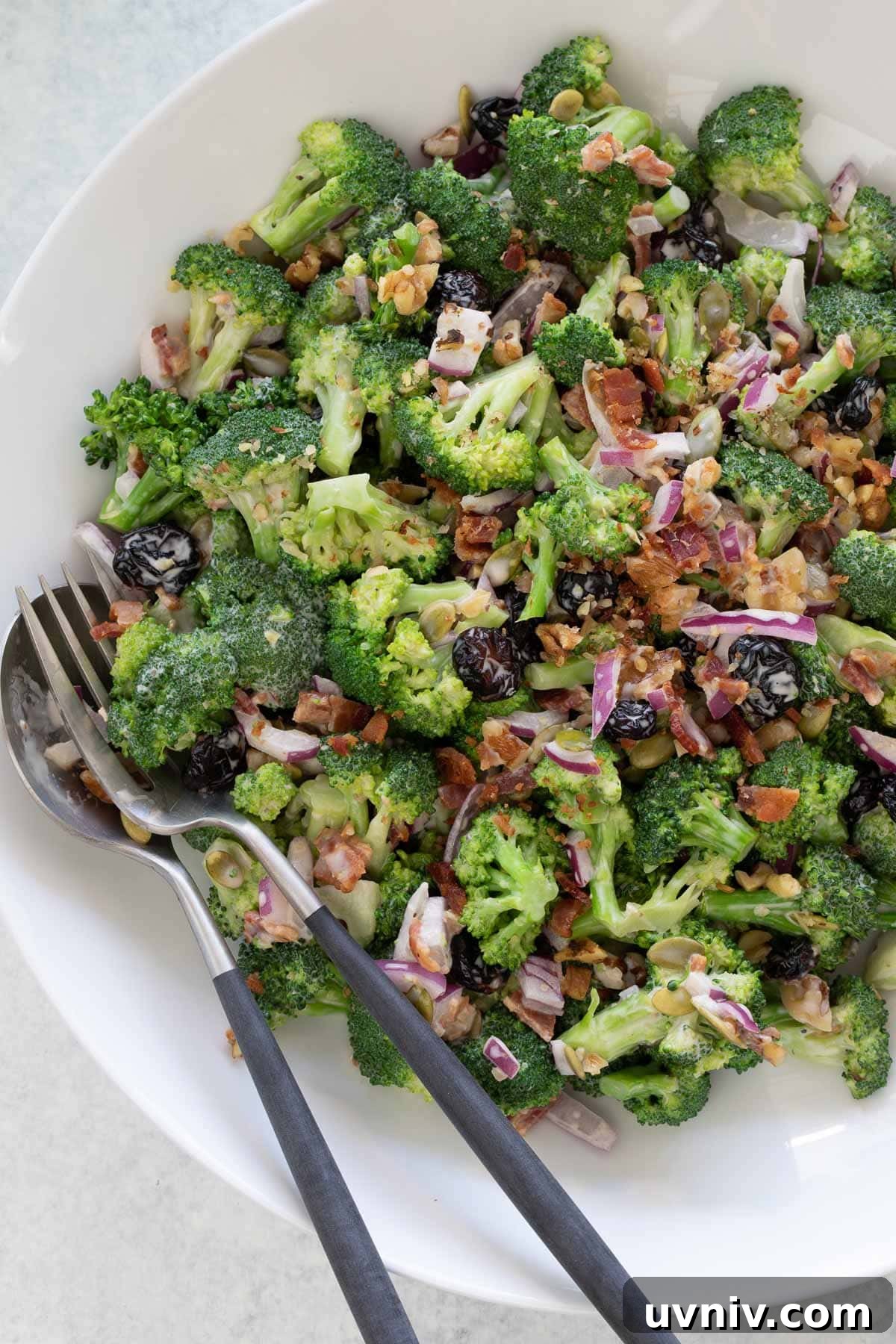 Vivid green broccoli salad with bacon, red onions, raisin, and walnuts in white bowl.