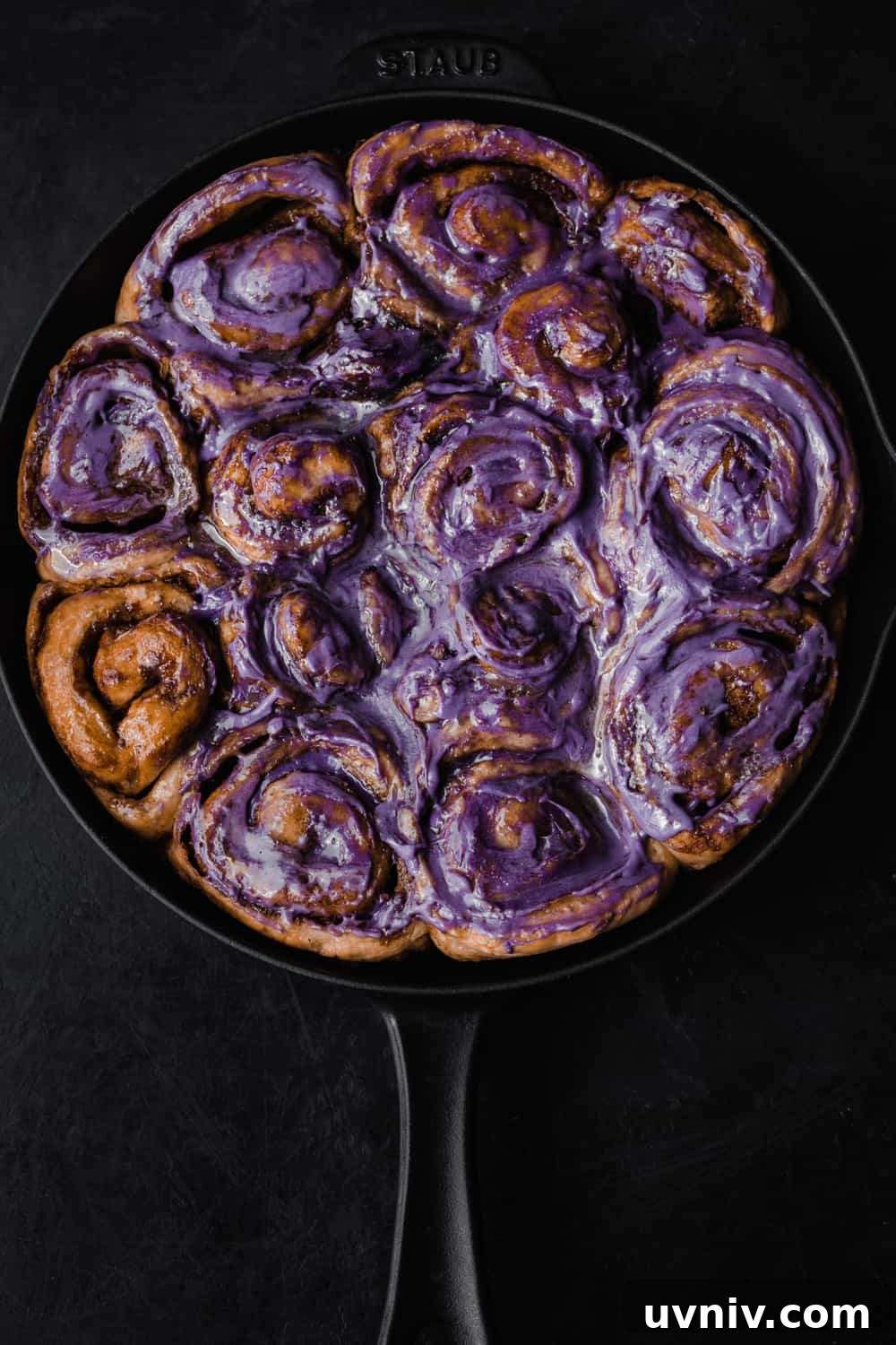 Cinnamon Rolls Made with Fluffy Challah Dough 6 Cinnamon rolls almost all frosted with a purple ube infused cream cheese icing; overhead shot on a black background.