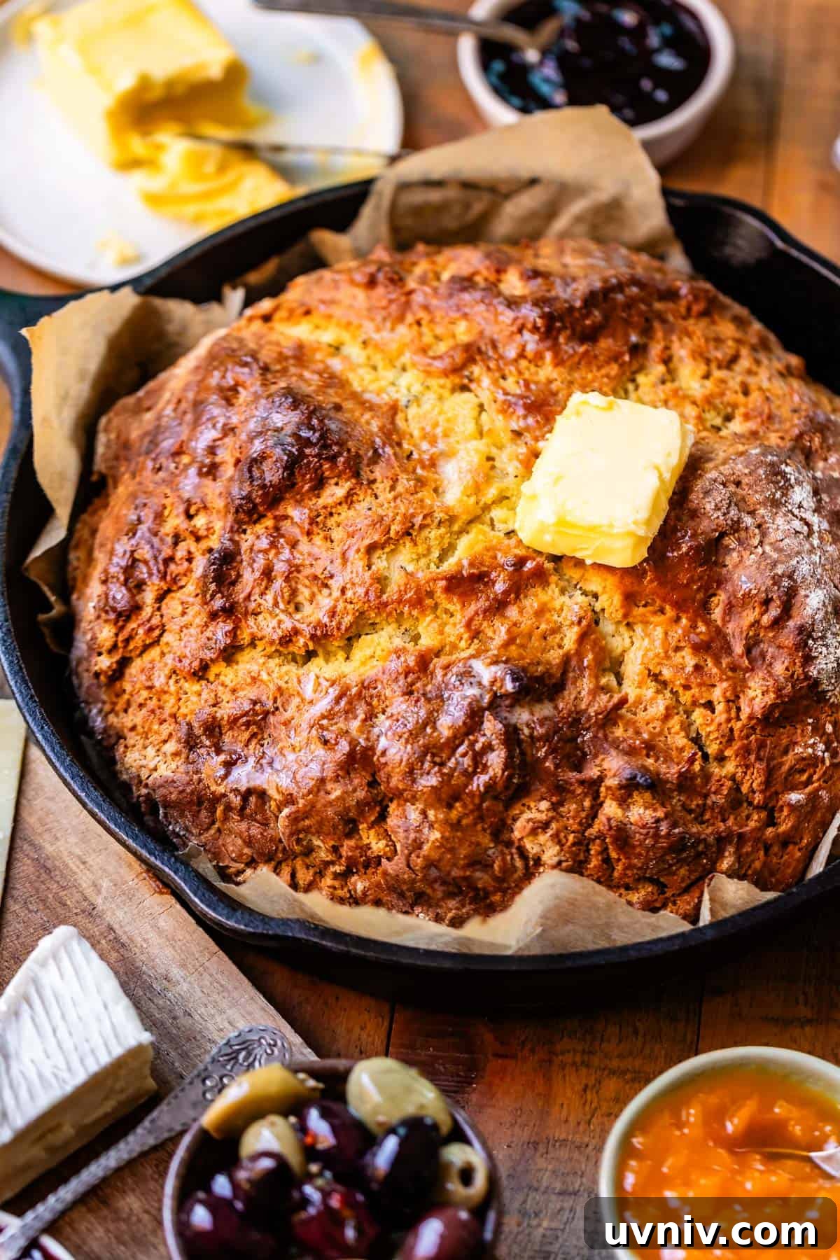 Authentic Irish Soda Bread Recipe You Can Make at Home 3 Baked round loaf of Irish soda bread with golden brown crusty top on parchment paper in cast iron skillet with toppings surrounding including butter, jam, and olives.