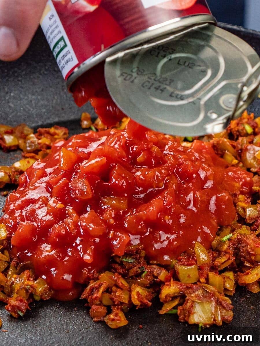 Spiced Brinjal Bhaji: Authentic Indian Eggplant Curry Recipe 7 Chopped tomatoes being poured into a frying pan on top of fried onions and spices