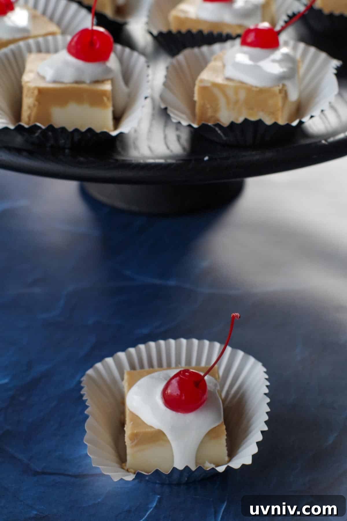 Root Beer Float Fudge Recipe: Creamy Soda-Infused Candy 20 a piece of root beer float fudge on a cupcake liner, in front of a black tray with more root beer float fudge pieces