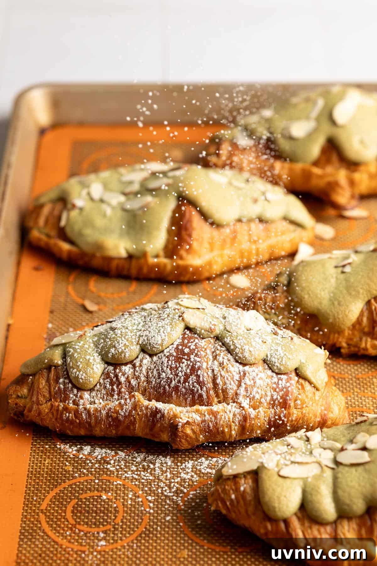 Matcha Almond Croissants Recipe: Flaky Green Tea Pastry Tutorial 2 Powdered sugar is being dusted on matcha almond croissants on a baking sheet.
