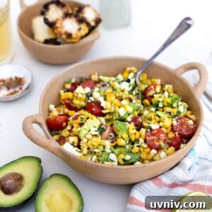 Grilled Corn and Avocado Salad with Lime Cilantro Dressing 13 A bowl of grilled corn avocado salad with cherry tomatoes, feta, and herbs sits on a white table. Nearby are a halved avocado, lime, grilled corn slices, a drink, and a vase with yellow flowers.