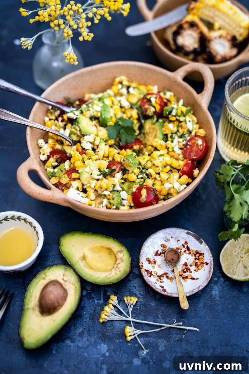 Grilled Corn and Avocado Salad with Lime Cilantro Dressing 3 A bowl of grilled corn avocado salad with tomatoes, feta, and herbs sits on a table with cut avocado, corn, lime, spices, a glass of lemonade, and yellow flowers nearby.