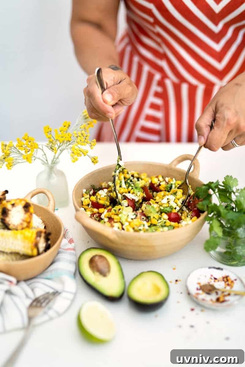 Grilled Corn and Avocado Salad with Lime Cilantro Dressing 4 A person in a red and white striped dress mixes a vibrant grilled corn avocado salad with tomatoes and herbs in a bowl. Nearby are fresh avocados, grilled corn, a lime half, herbs, and a vase with yellow flowers.