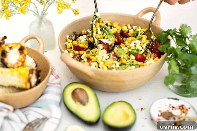Grilled Corn and Avocado Salad with Lime Cilantro Dressing 8 A person in a red and white striped dress mixes a vibrant grilled corn avocado salad with tomatoes and herbs in a bowl. Nearby are fresh avocados, grilled corn, a lime half, herbs, and a vase with yellow flowers.