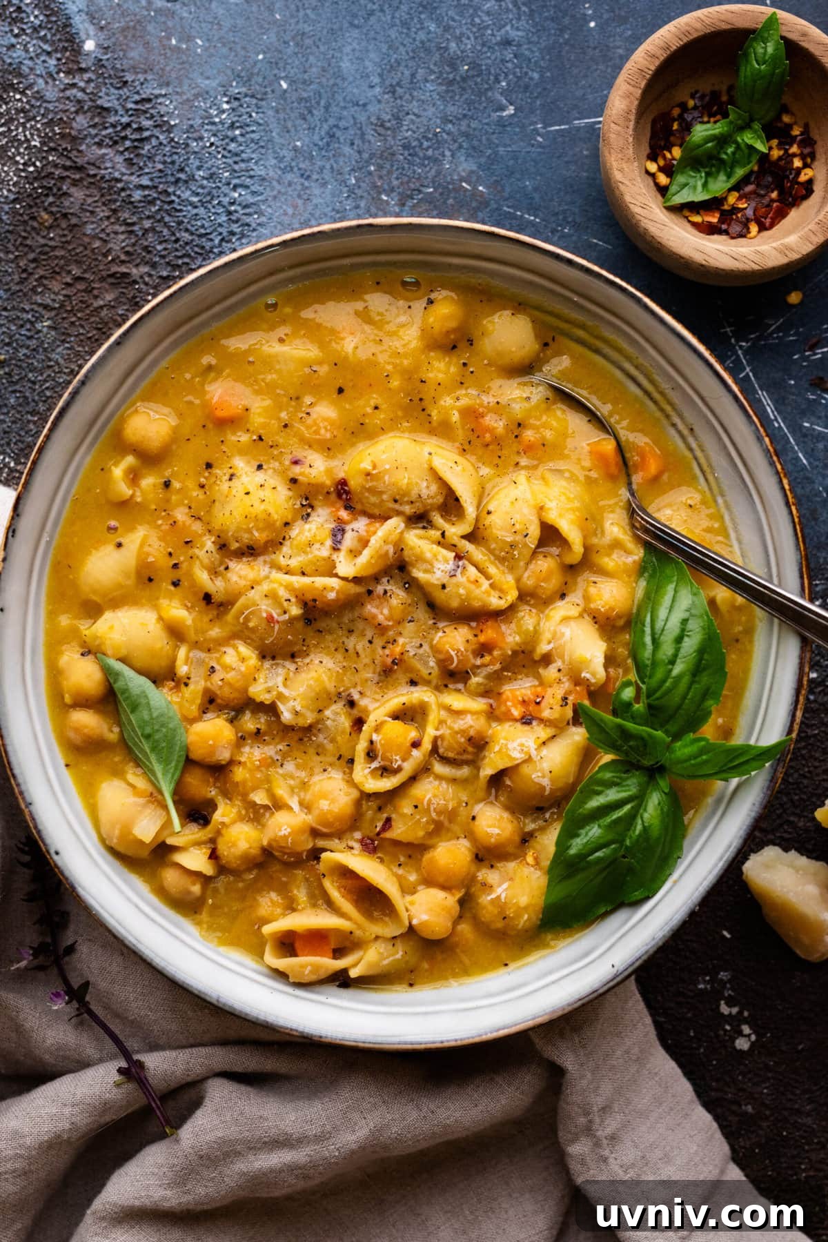Pasta e Ceci: Classic Italian Pasta with Chickpeas Recipe 2 A bowl filled with pasta e ceci soup and fresh herbs on top. The bowl is sitting on a dark background with a napkin and small bowl of red pepper flakes arranged around it.