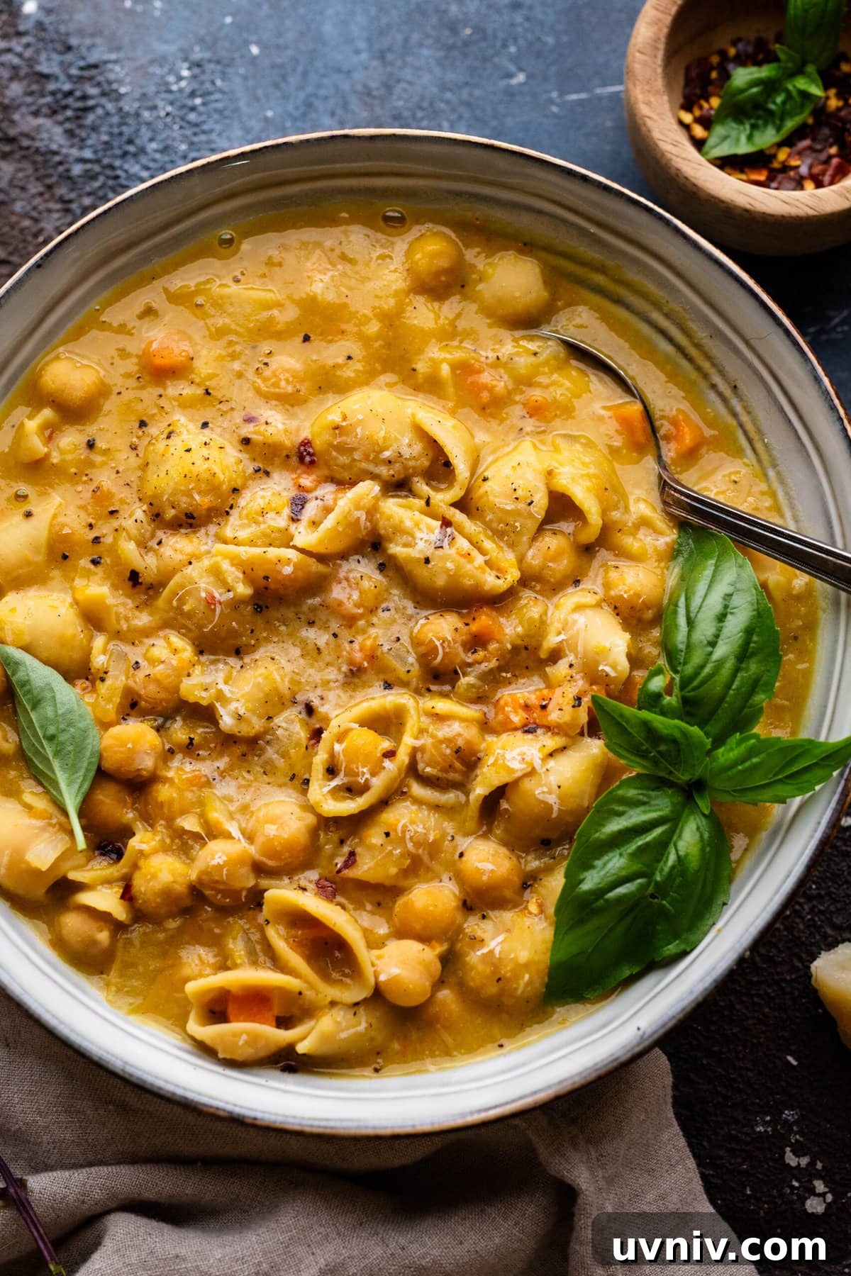 Pasta e Ceci: Classic Italian Pasta with Chickpeas Recipe 7 A bowl filled with pasta e ceci soup and fresh herbs on top. The bowl is sitting on a dark background with a napkin and small bowl of red pepper flakes arranged around it.