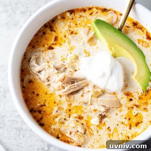 Low-Carb White Chicken Chili Recipe for Keto Diet 17 overhead view of bowl of keto white chicken chili in white serving bowl and gold serving spoon and garnished with slice of avocado and sour cream
