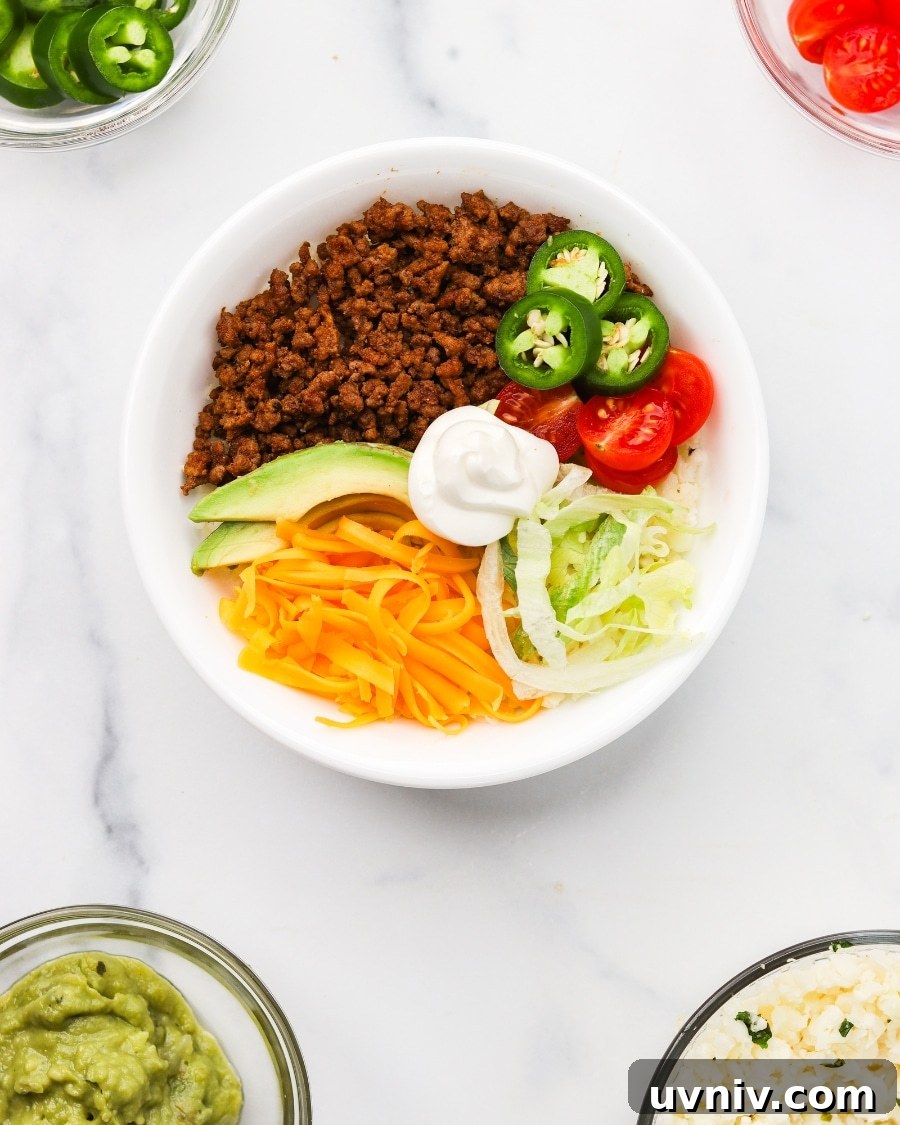 keto taco bowl assembled with bowls of toppings nearby including guac, cherry tomatoes, cauliflower rice, and jalapenos