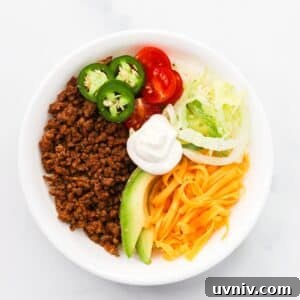 High protein keto taco bowl with ground beef, shredded cheddar, avocado, cherry tomatoes, and jalapeños in a white bowl
