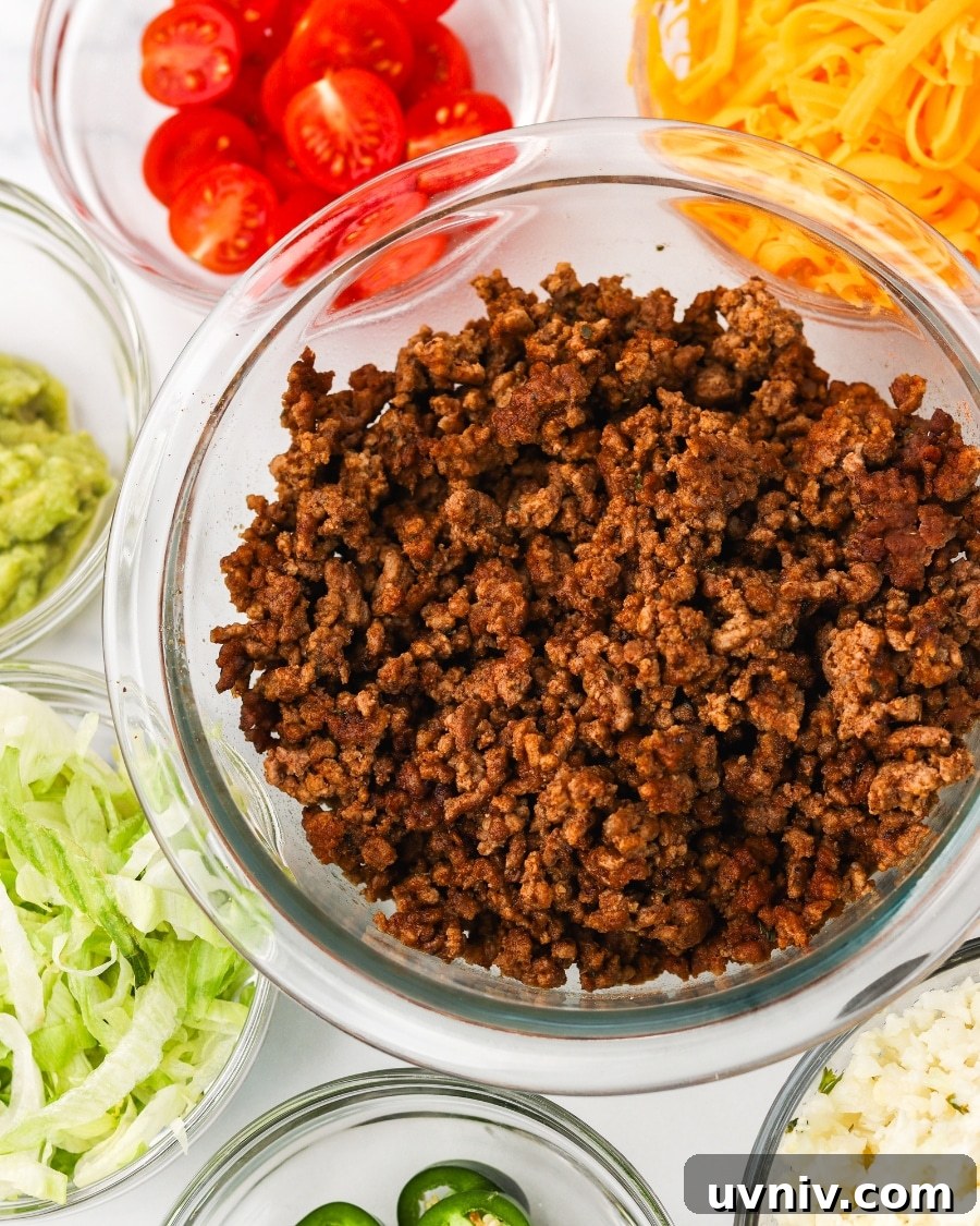 All ingredients ready to be assembled for keto taco bowls including ground beef, lettuce, tomato, shredded cheddar cheese, cauliflower rice, guacamole, and sliced jalapenos