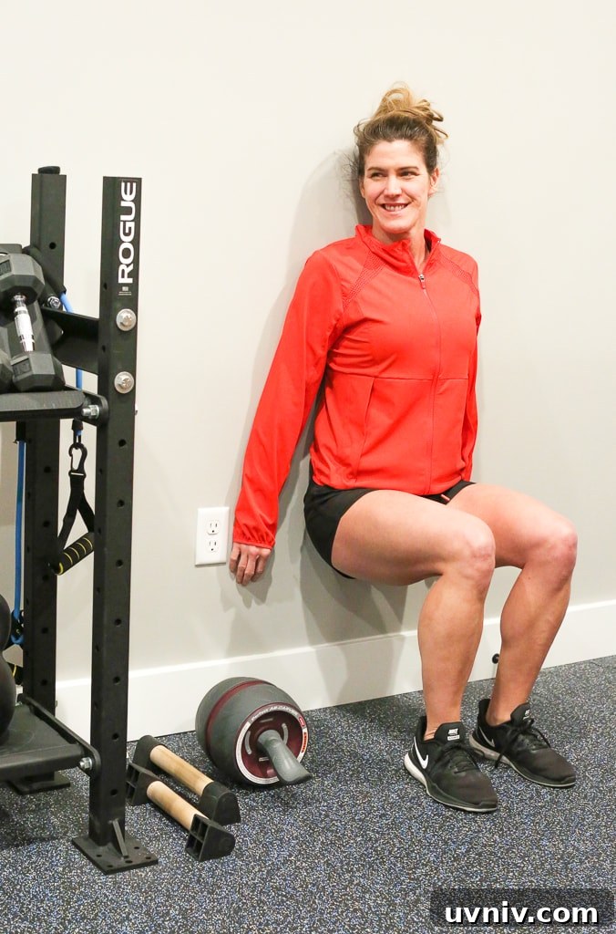 Effective At-Home Leg Workout Routine for Strength and Tone 3 Maryea Flaherty with her back against the wall and legs at a 90 degree angle, performing wall sit exercise in a red jacket and black shorts