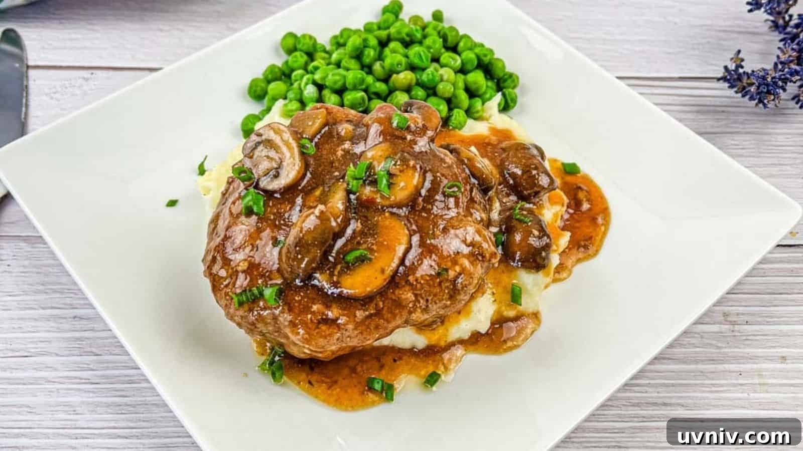A white plate with Salisbury steak topped with mushroom gravy on mashed potatoes, accompanied by a side of green peas.