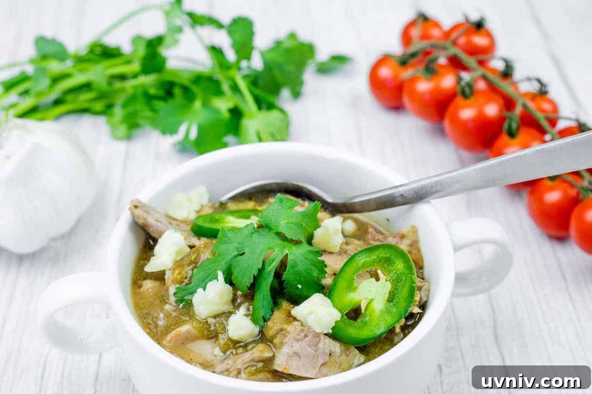 Bowl of Slow Cooker Pork Chile Verde with toppings and a spoon on a white wooden table.