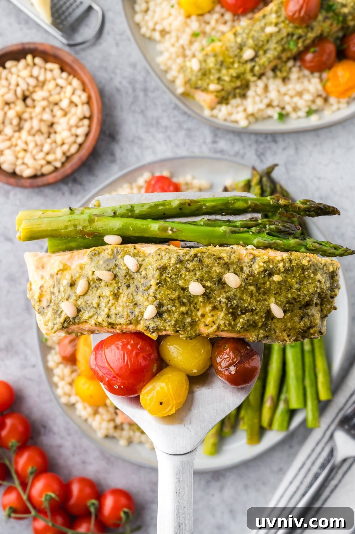 One-Pan Salmon and Roasted Asparagus Recipe 12 A spatula holds pesto salmon with asparagus and cherry tomatoes over a plate of couscous and vegetables.