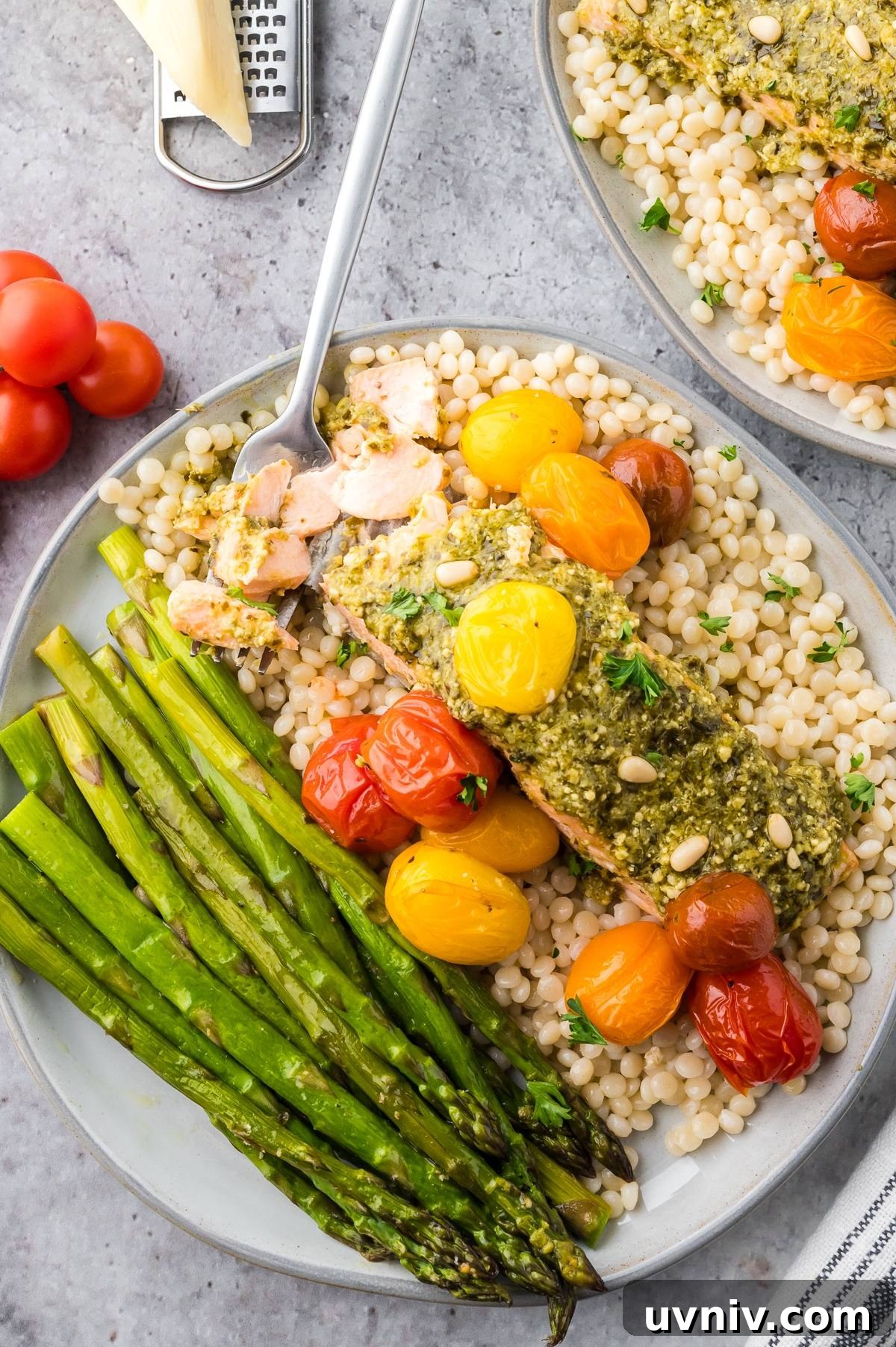 One-Pan Salmon and Roasted Asparagus Recipe 13 Plate of salmon with pesto, roasted tomatoes, asparagus, and pearl couscous on a gray surface.