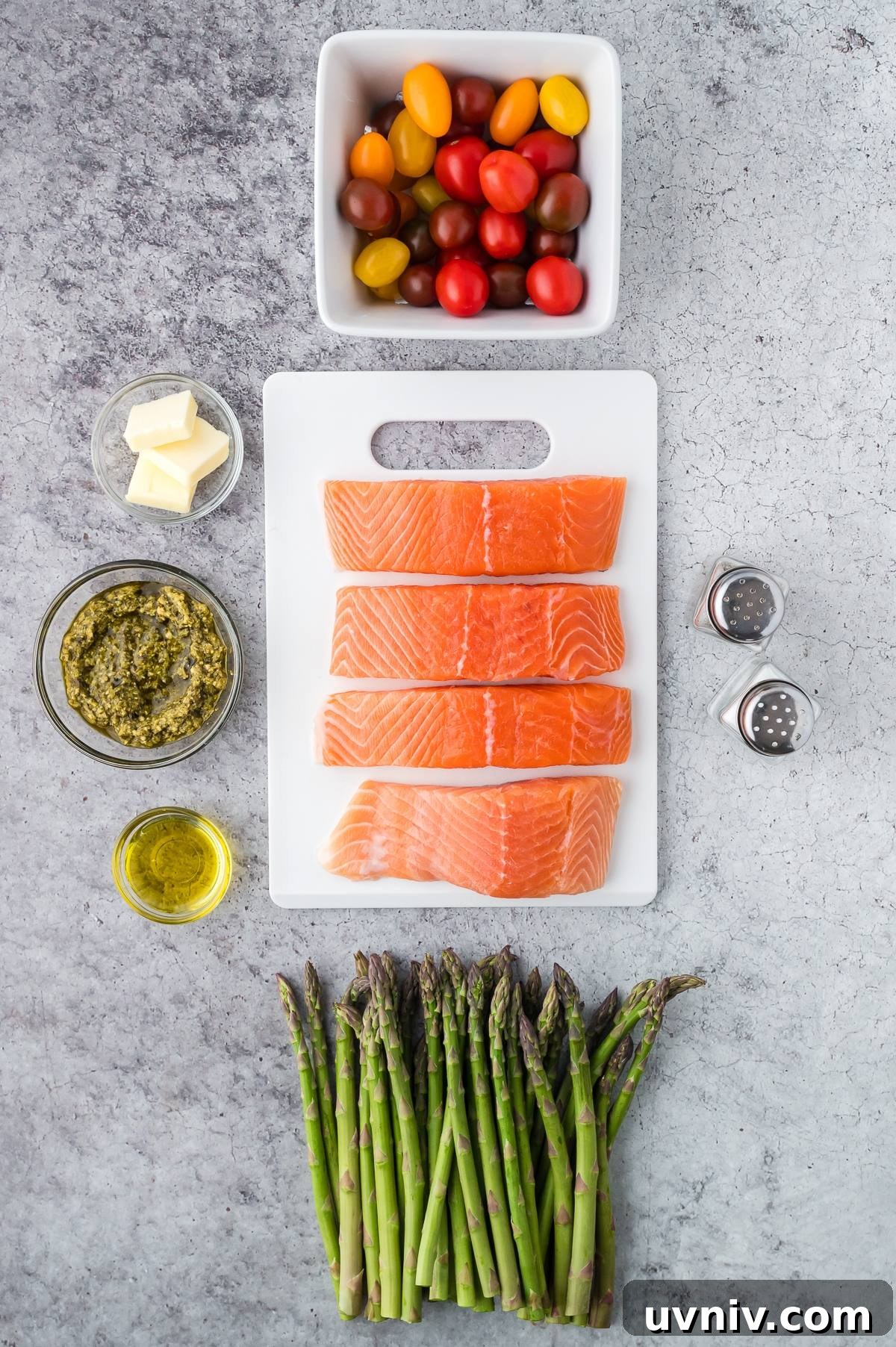 One-Pan Salmon and Roasted Asparagus Recipe 7 Four raw salmon fillets, asparagus, tomatoes, pesto, butter, olive oil, salt, and pepper on a gray surface.