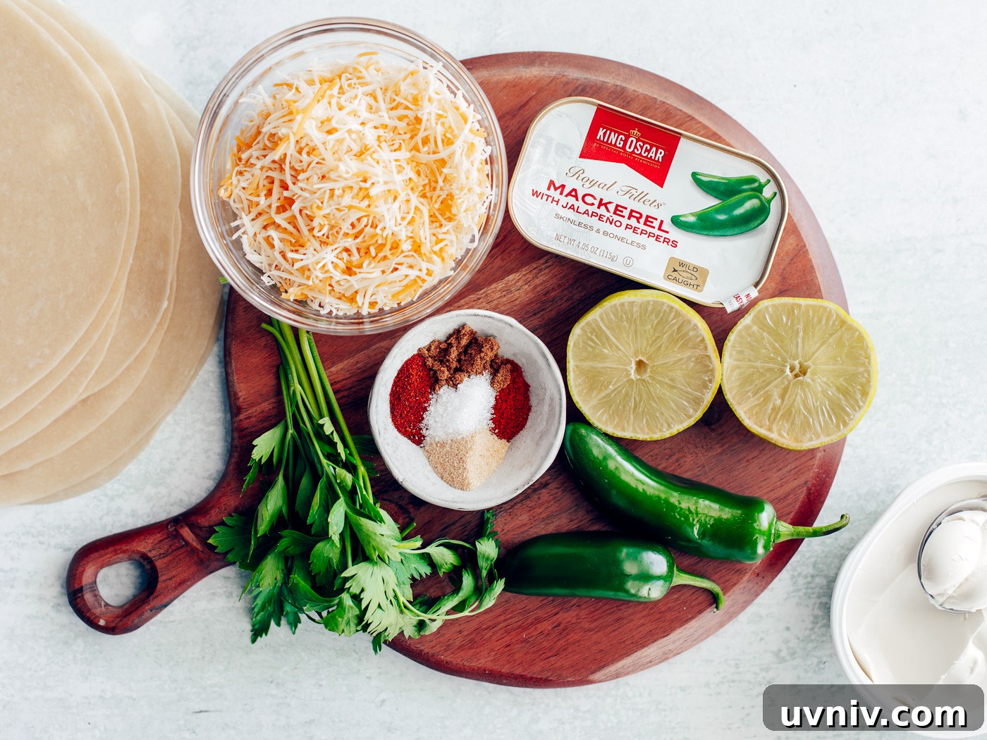 Round serving board with tin of mackerel, bowls of grated cheese and spice mix, jalapenos, limes, and parsley.