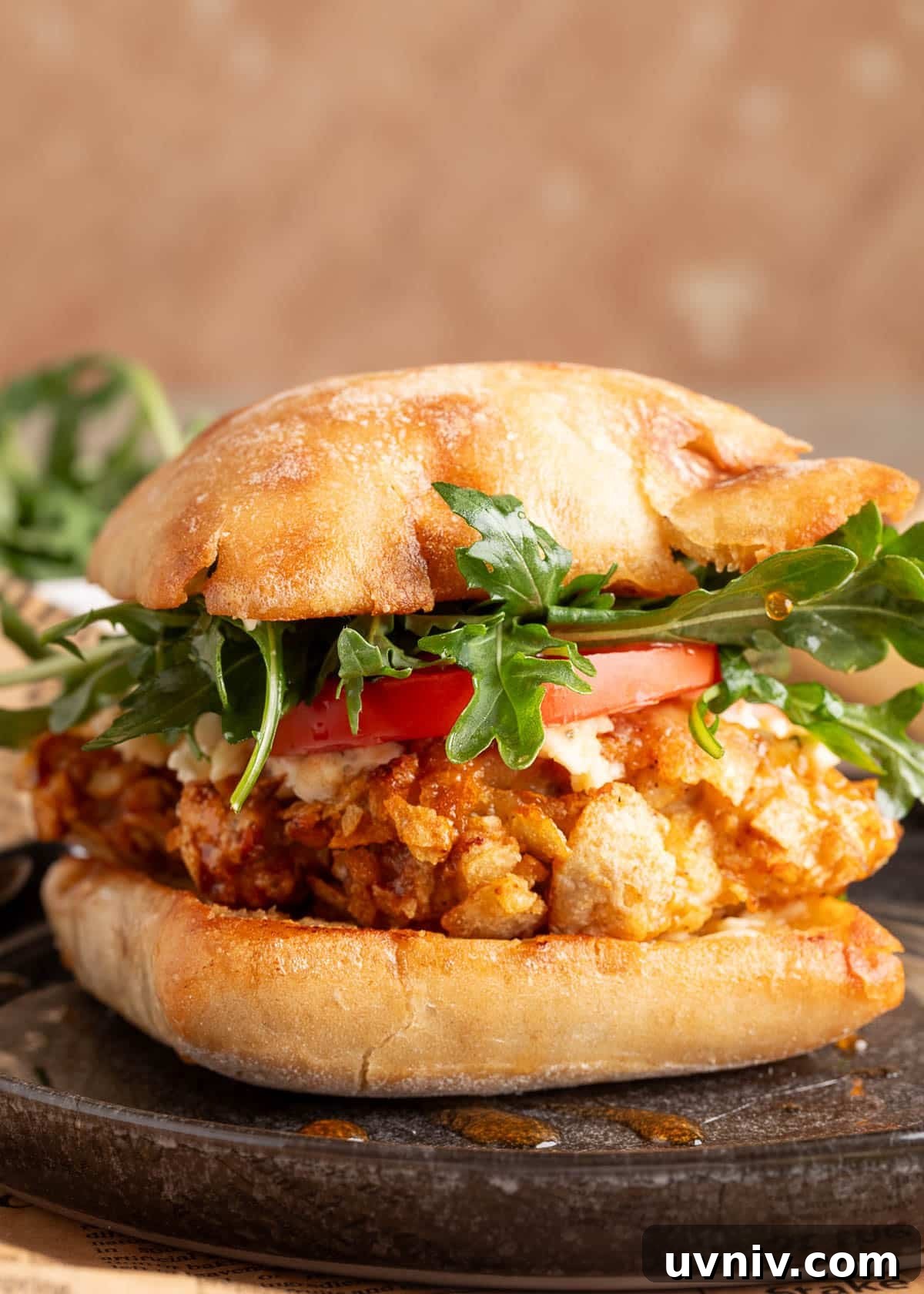 Close-up of a crispy air fryer chicken sandwich on a toasted ciabatta roll, layered with potato chip-crusted chicken, sliced tomato, arugula, and crumbled blue cheese, with hot honey drizzled underneath.