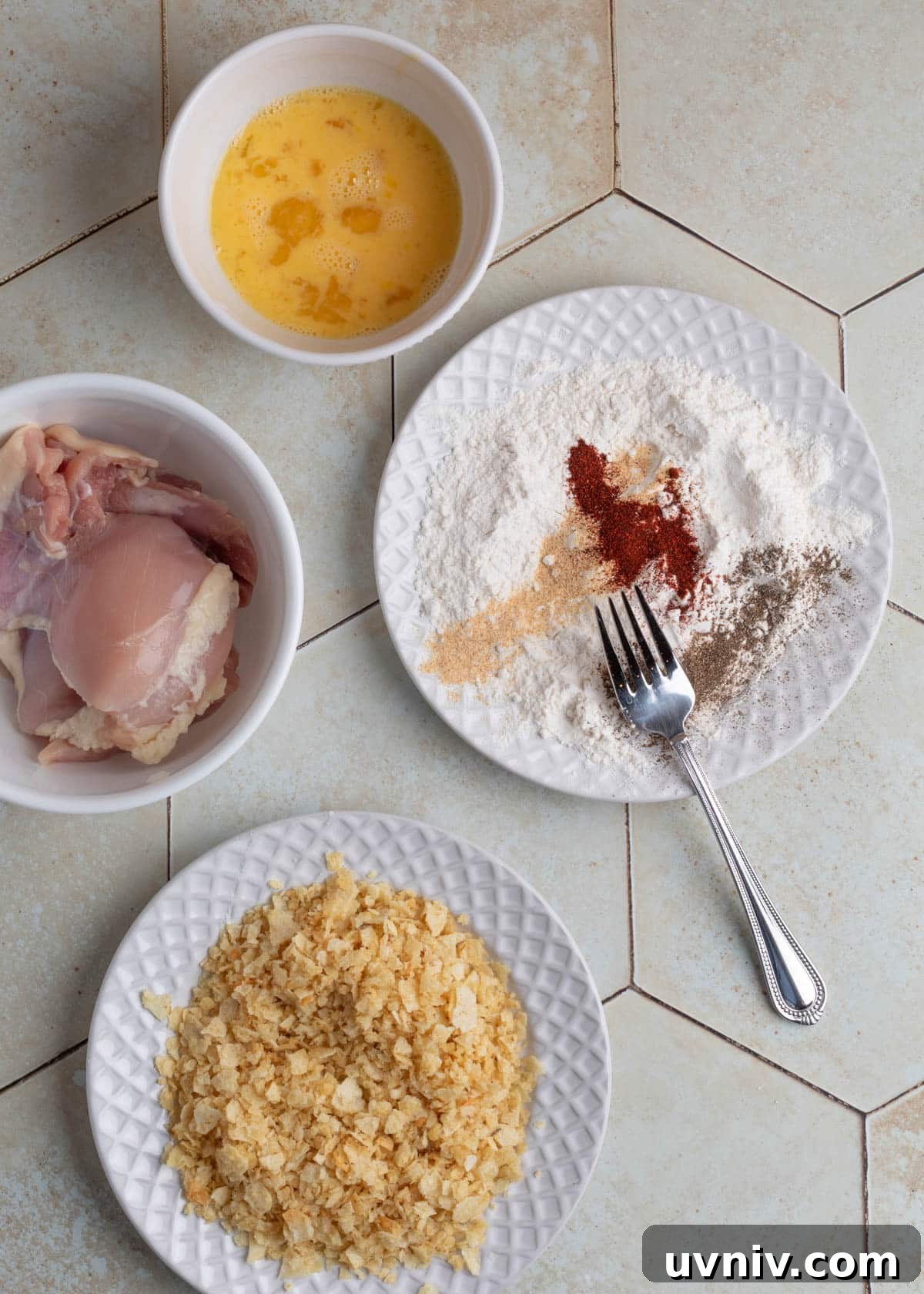 Overhead view of prep bowls with raw chicken, egg wash, crushed potato chips, and a white plate of flour with paprika, garlic powder, onion powder, pepper, and a fork ready to mix.