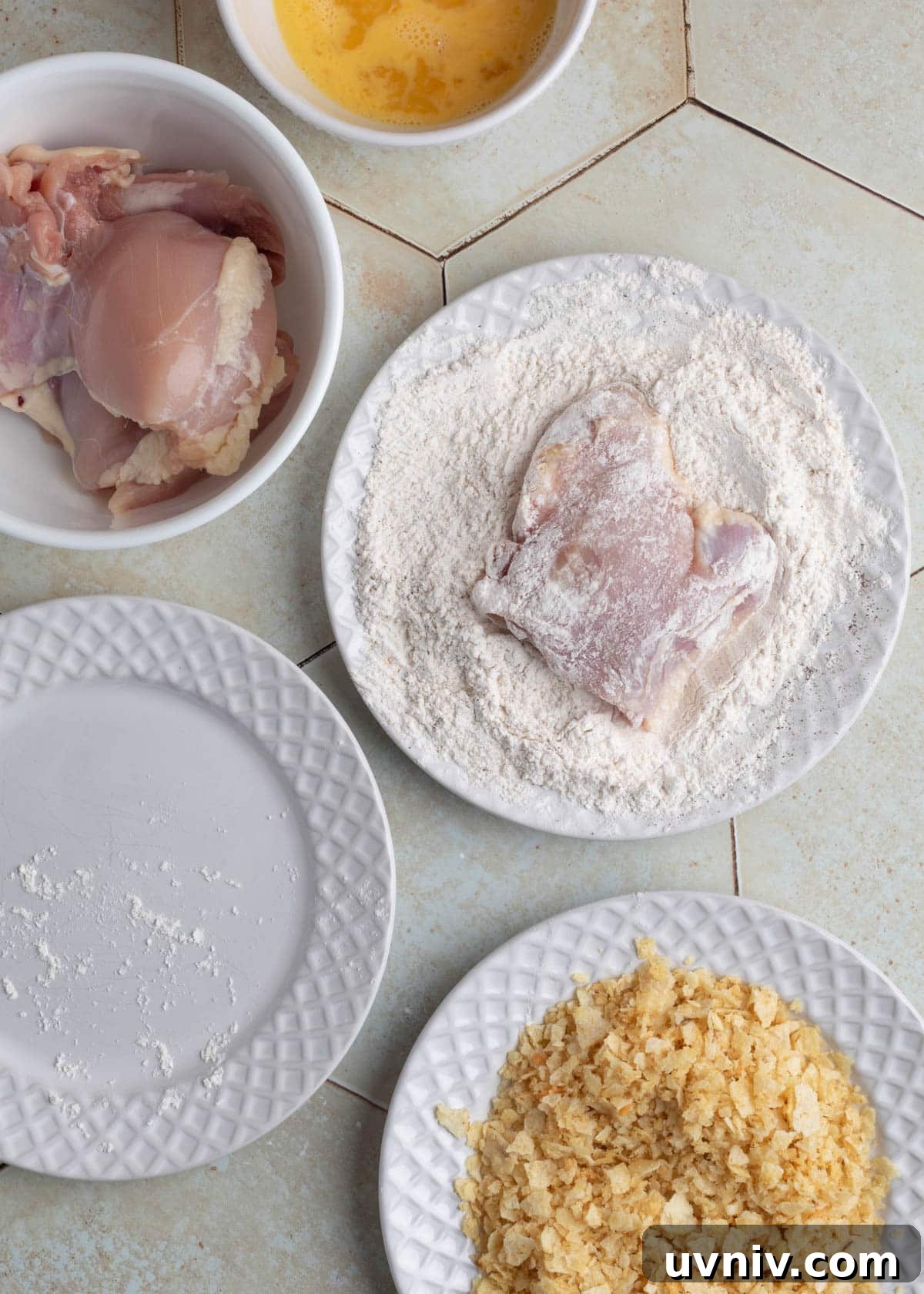 Raw chicken thigh dredged in seasoned flour, resting on a white plate, next to bowls of egg wash, raw chicken thighs, and crushed chips.