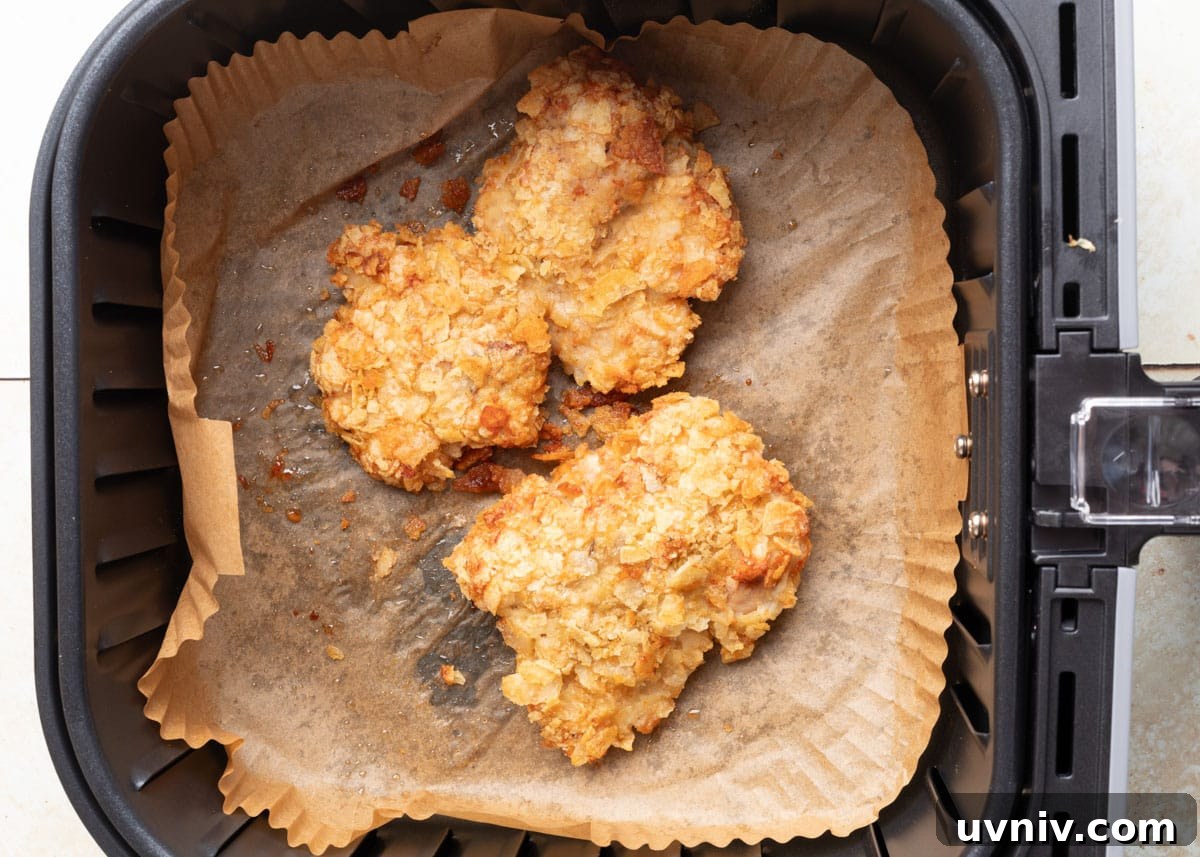 Two crispy potato chip–crusted chicken thighs in an air fryer basket lined with parchment paper, freshly cooked and golden brown.
