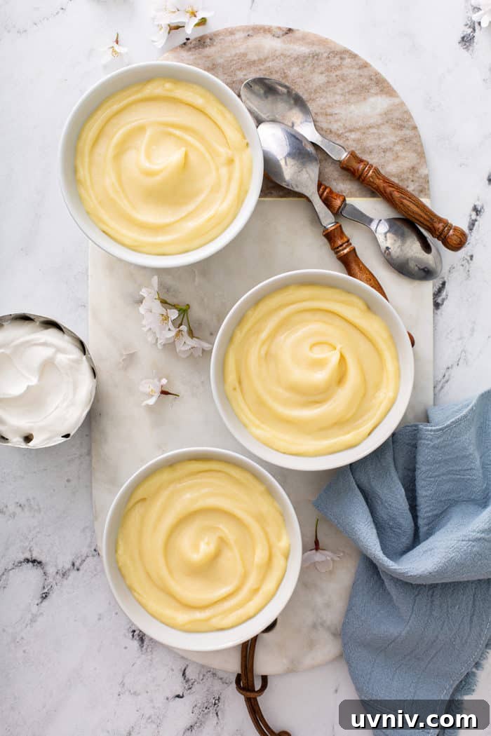 Overhead view of three white bowls of homemade vanilla pudding on a marble board, surrounded by spoons and homemade whipped cream