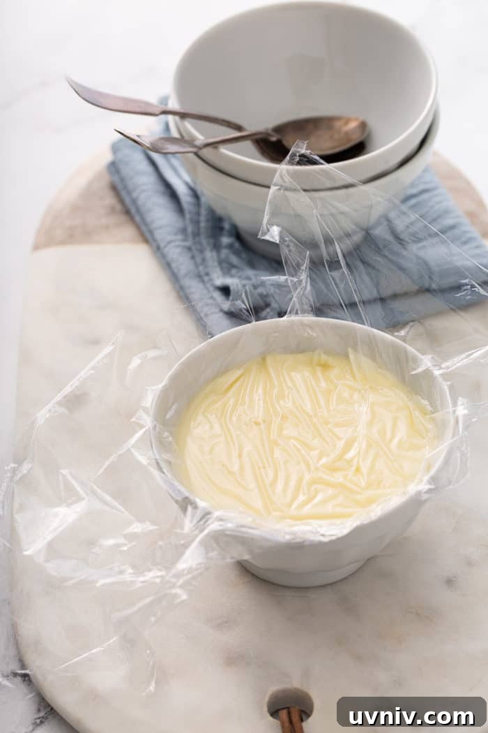 Small white bowl full of homemade vanilla pudding with plastic wrap pressed onto the top of the pudding