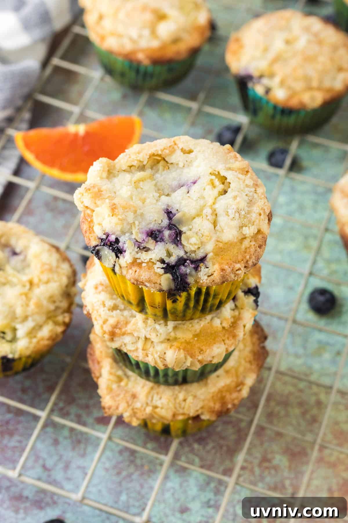Moist Blueberry Orange Muffins with Zesty Citrus Glaze 2 Three blueberry orange muffins with crumb topping stacked on a cooling rack, with loose blueberries and a slice of orange nearby.