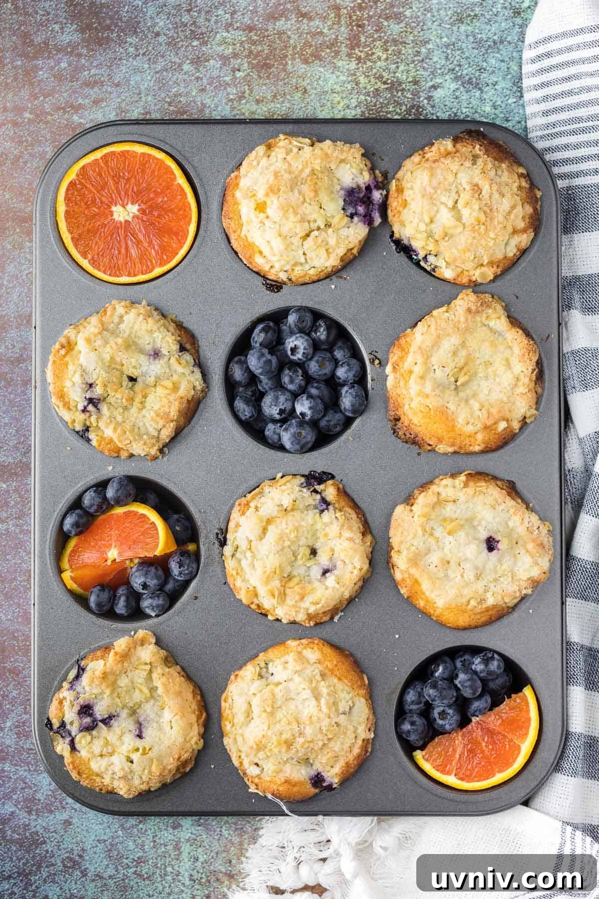 Moist Blueberry Orange Muffins with Zesty Citrus Glaze 4 A muffin tin with nine baked blueberry muffins, three empty cups filled with fresh blueberries and orange slices, on a textured surface with a striped towel nearby.