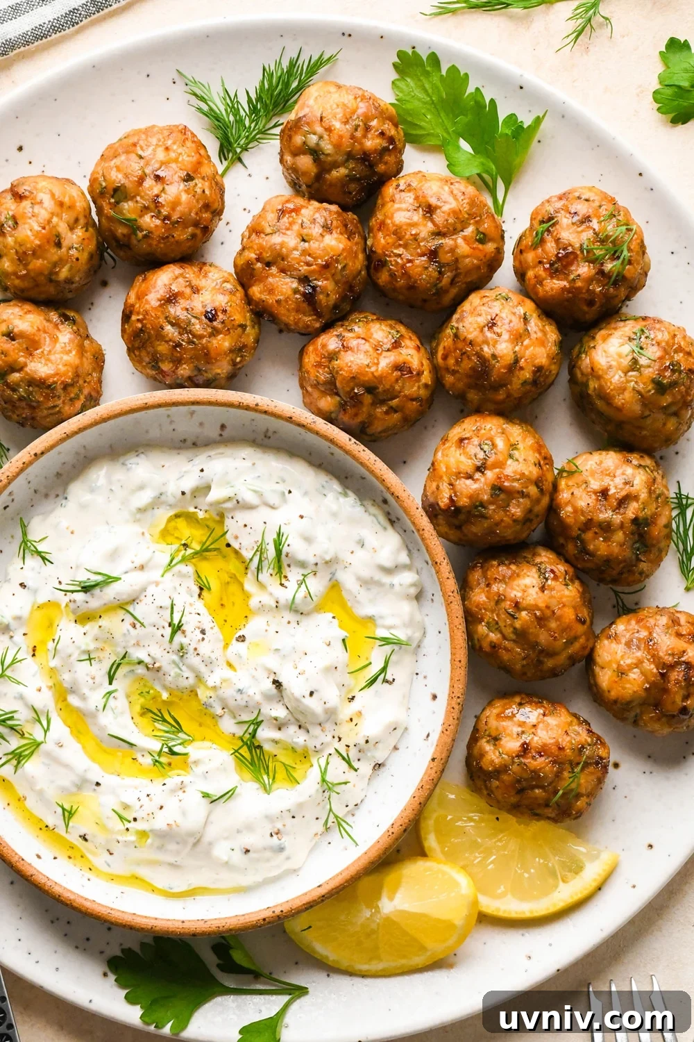 Baked Greek chicken meatballs on a serving platter, next to a small dish of tzatziki, and garnished with a few sprigs of fresh dill and fresh parsley.