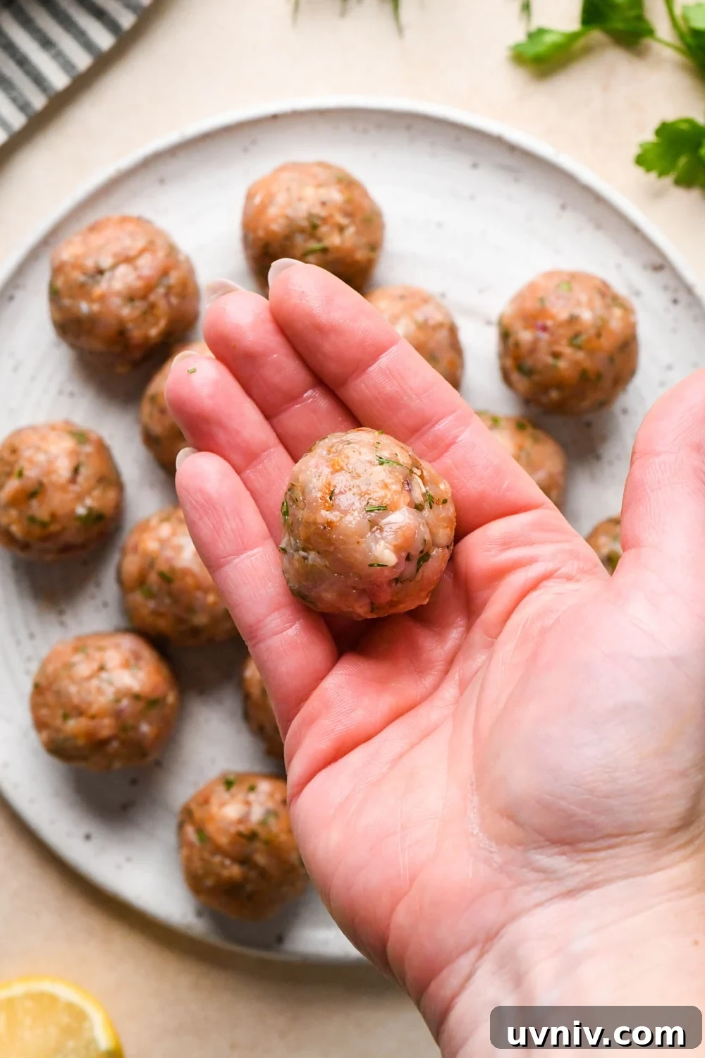 How to make Greek chicken meatballs: A hand that has been wet with water showing a meatball after rolling it to shape over a plate of meatballs that have already been shaped.