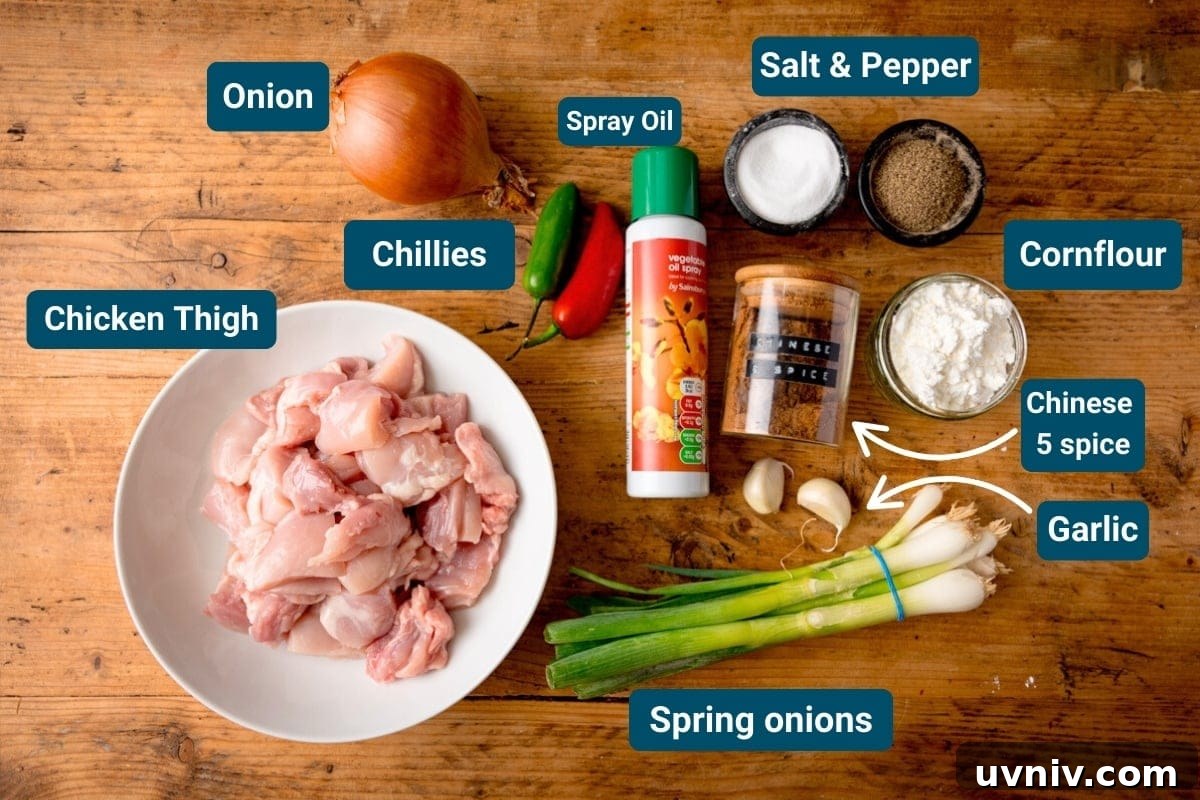 Crispy Salt and Pepper Chicken Made in the Air Fryer 4 Labelled ingredients for making air fryer salt and pepper chicken on a wooden surface including: chopped chicken thigh, onion, chillies, spring onions, garlic, spray oil, salt, pepper, cornflour and Chinese five spice.