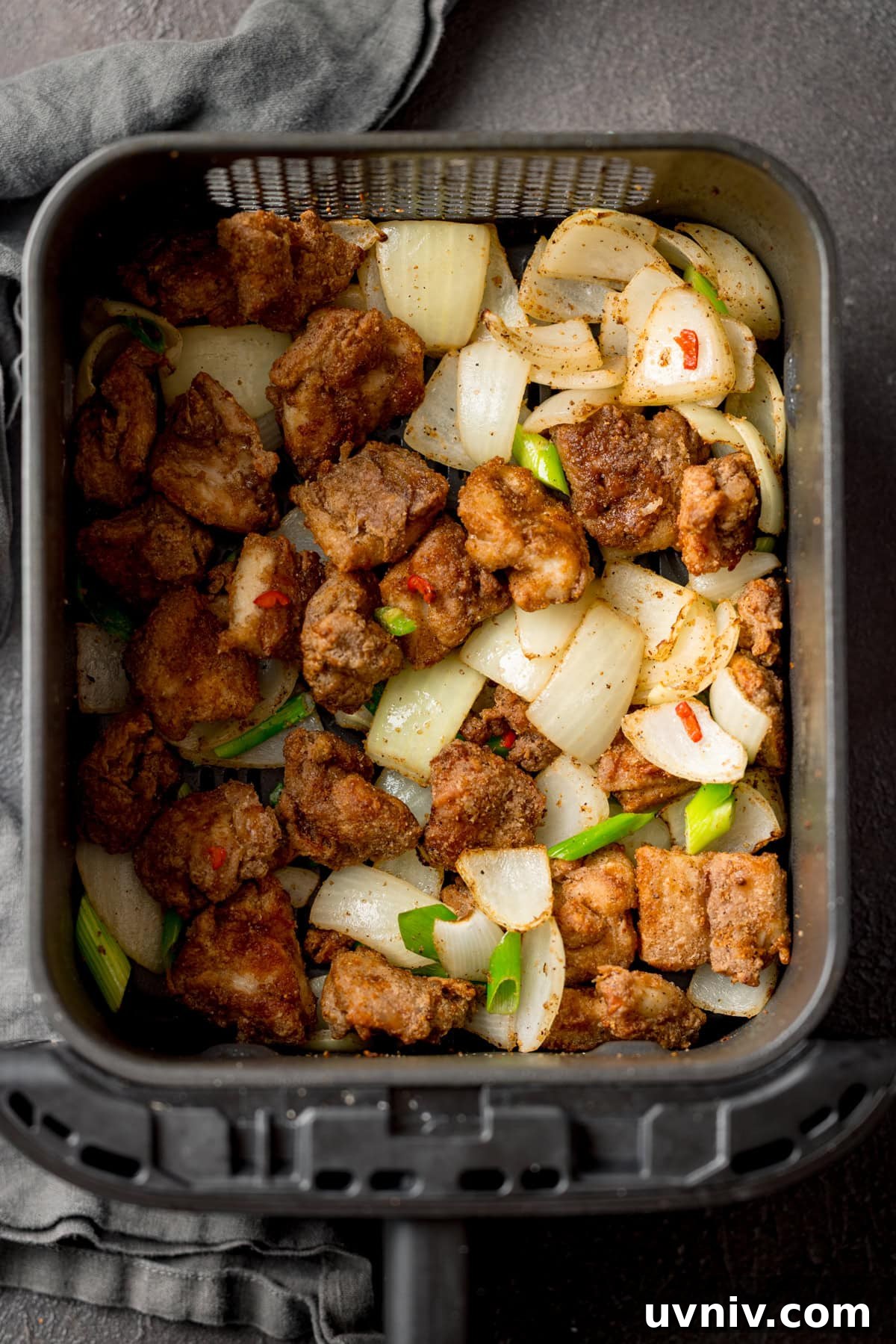 Crispy Salt and Pepper Chicken Made in the Air Fryer 5 Salt and Pepper Chicken pieces with chopped onion, scallions and chillies in an air fryer basket, seen from above.