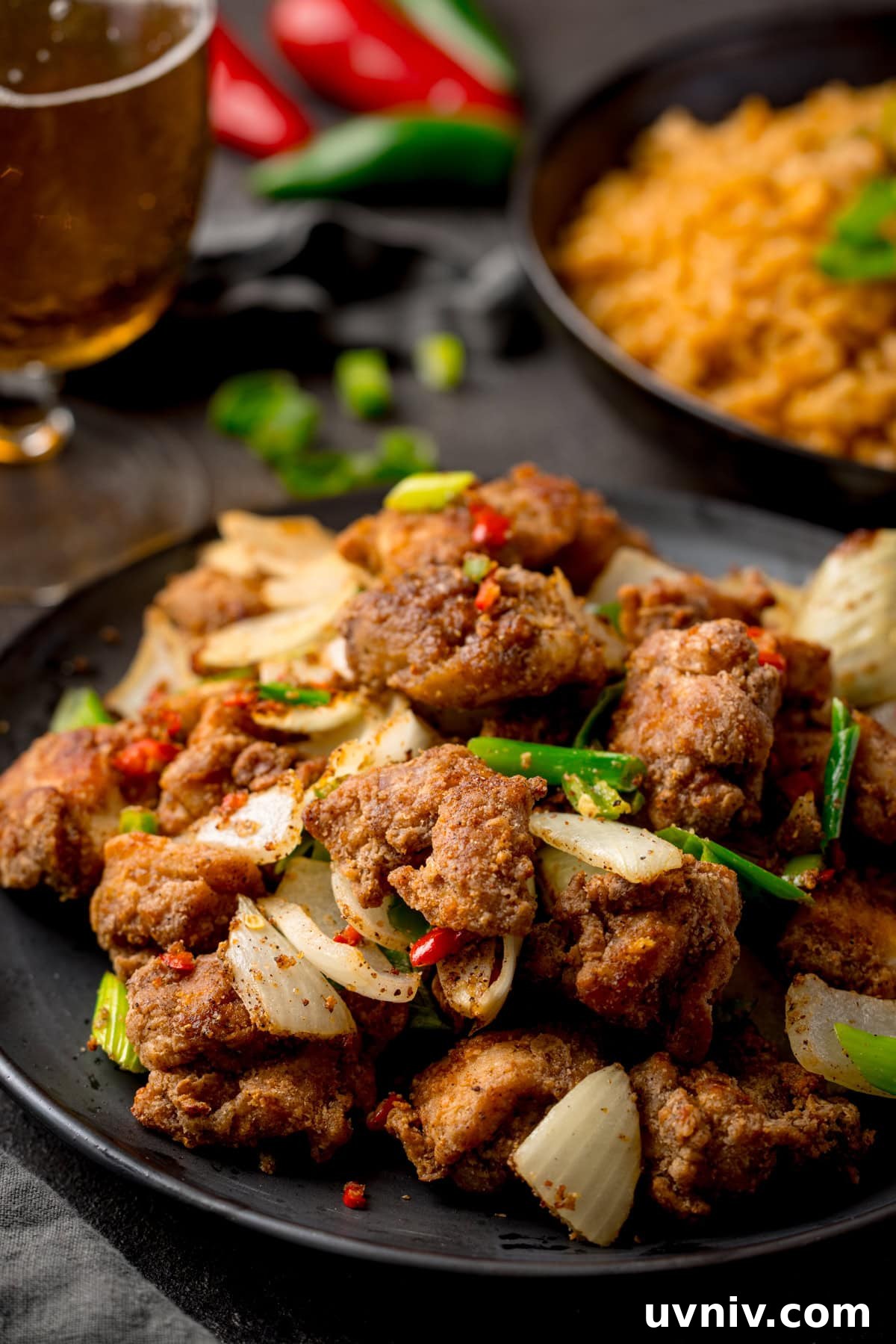 Crispy Salt and Pepper Chicken Made in the Air Fryer 8 Tall image of a plate of crispy Salt and Pepper Chicken pieces with onions, spring onions, and red chillies on a dark background. There is a plate of rice, a glass of beer and some fresh chillies partially visible in the background.