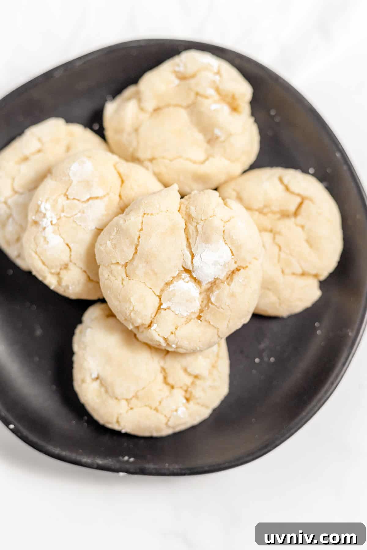 A black plate holds a stack of round, gooey cookies, their cracked surfaces dusted with powdered sugar on a pristine white surface.