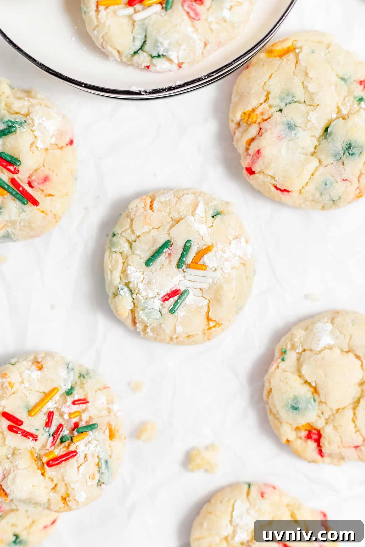 Gooey butter cookies adorned with vibrant sprinkles rest on crinkled parchment paper, with one temptingly perched on a plate.