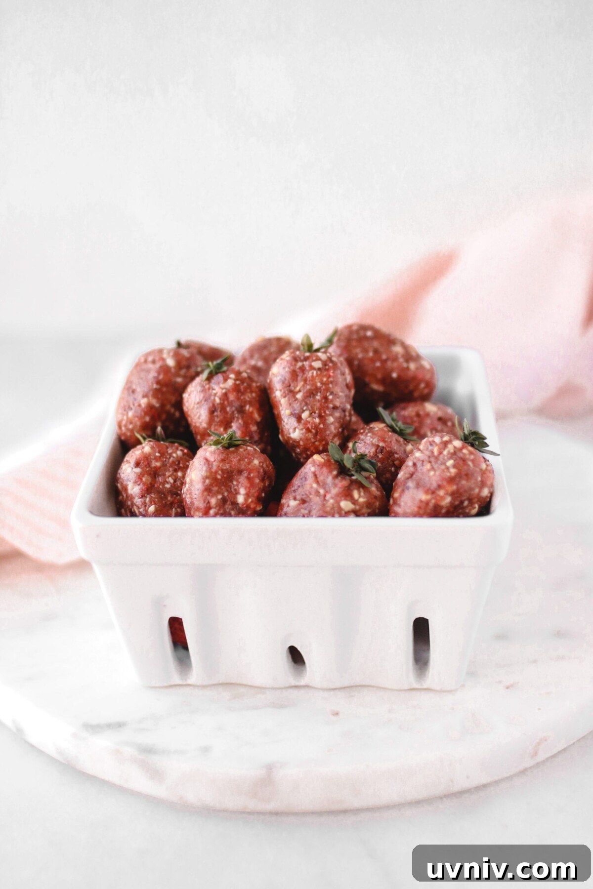 Strawberry Bliss Balls Recipe: No-Bake Energy Bites with Oats 3 A white ceramic container filled with strawberry-shaped energy bites made of nuts, seeds, and dried fruits. The bites are arranged to resemble strawberries with stems, placed on a round marble surface. A pink cloth is in the background.