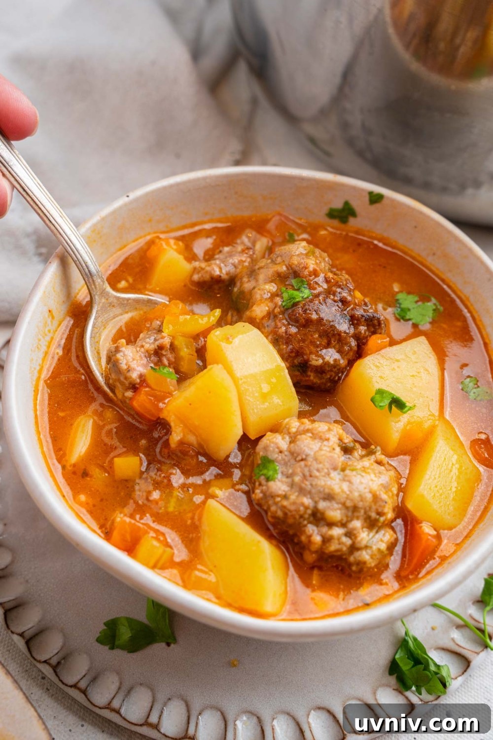 A spoon taking a bite of cajun meatball stew from a white ceramic bowl garnished with fresh parsley. The bowl is on a ceramic plate.