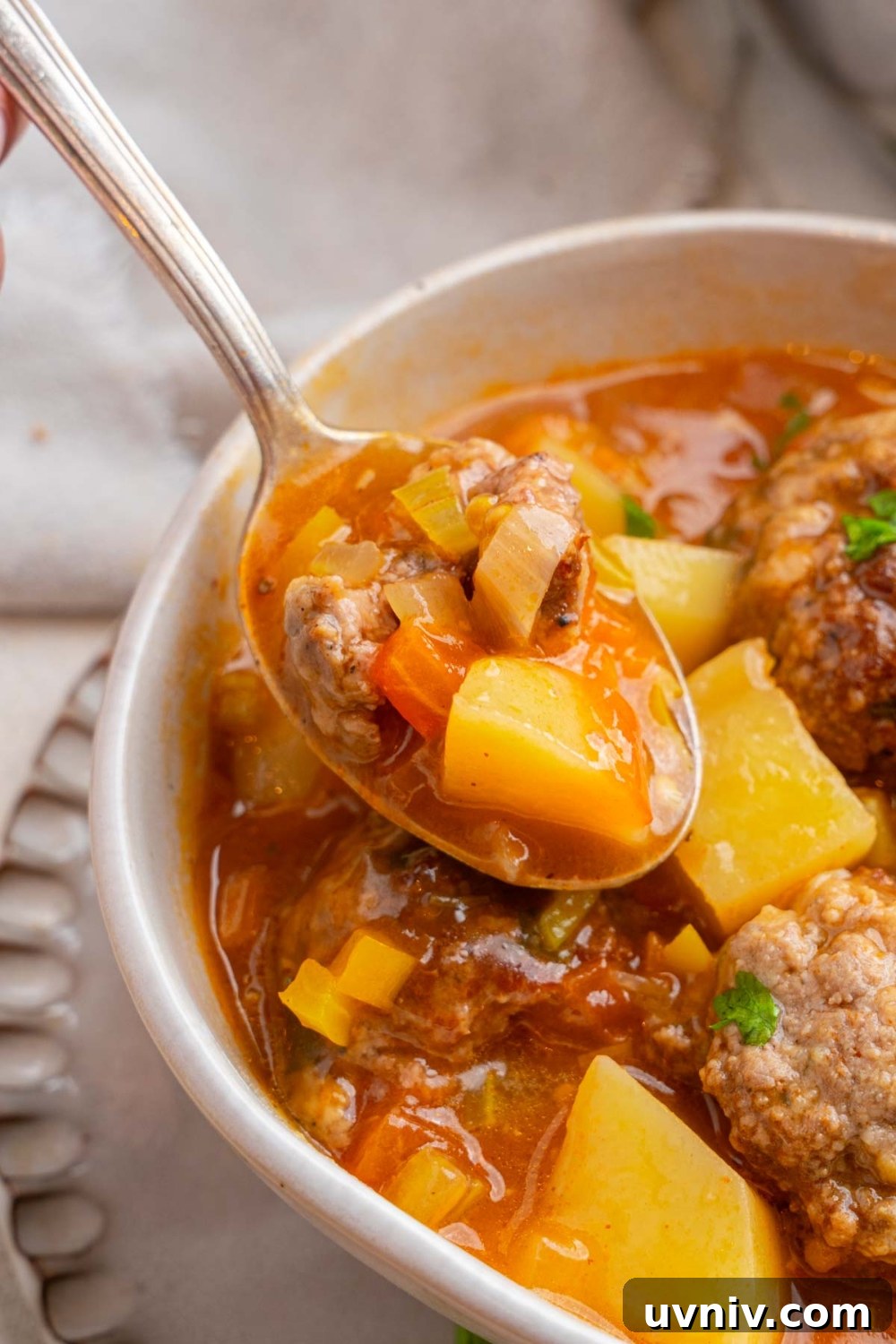 A spoon taking a bite of cajun meatball stew from a white ceramic bowl garnished with fresh parsley. The bowl is on a ceramic plate.