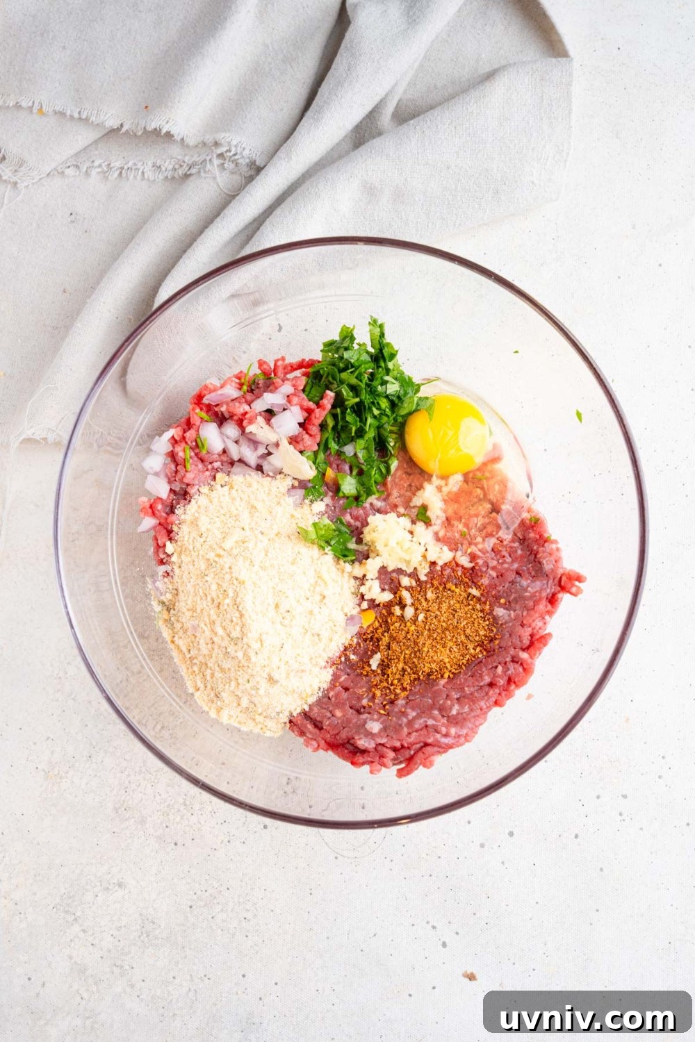 A glass bowl with ingredients to make cajun meatballs including ground beef, bread crumbs, parsley, egg, and seasonings. The bowl is on a white counter with a white cloth napkin.