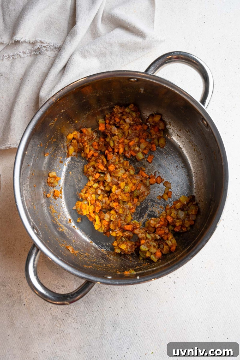 A large stainless steel pot with a roux sauteeing with diced vegetables. The pot is on a white counter with a white cloth napkin.