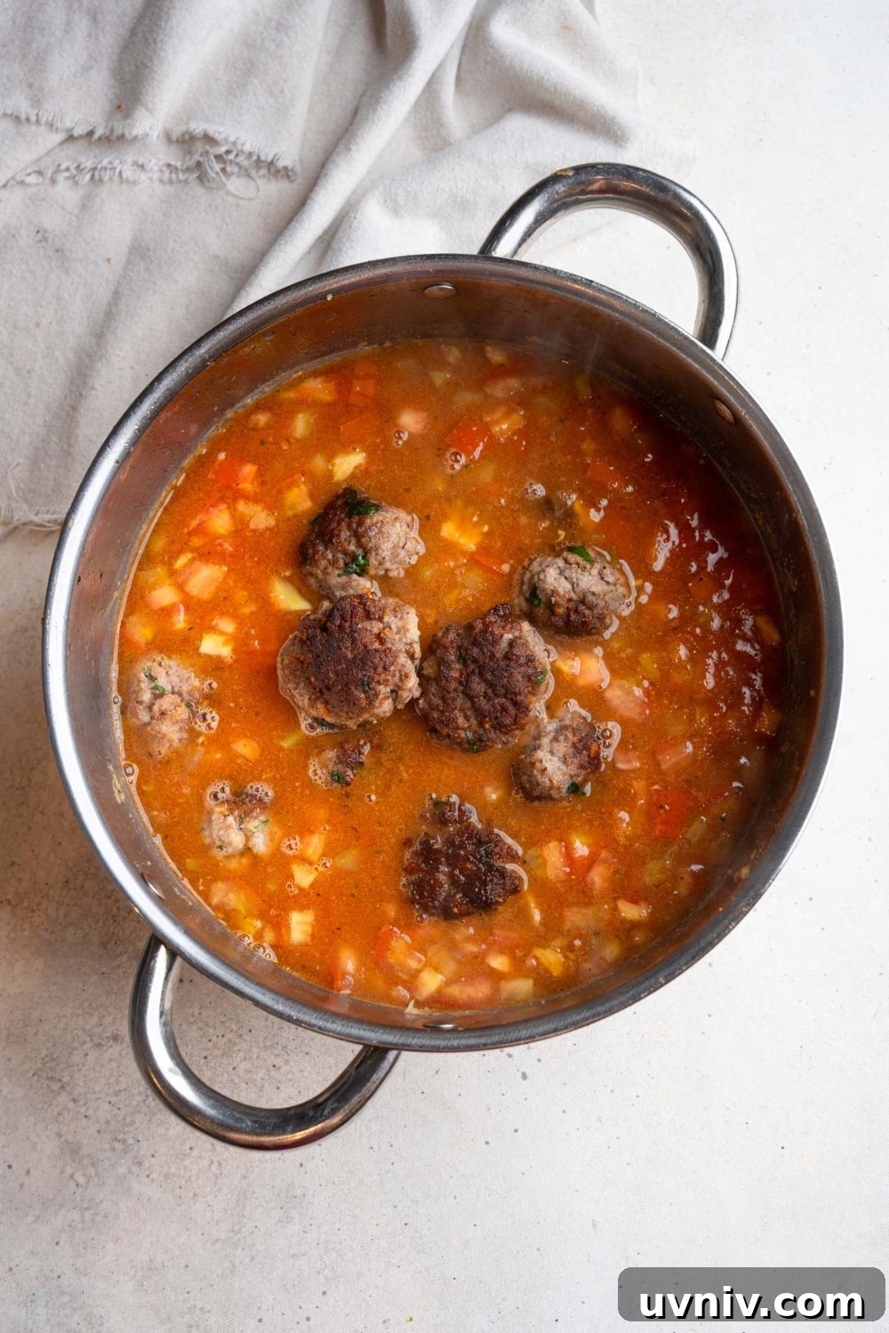 A large stainless steel pot with a cooked stew base and several meatballs floating on top. The pot is on a white counter with a white cloth napkin.