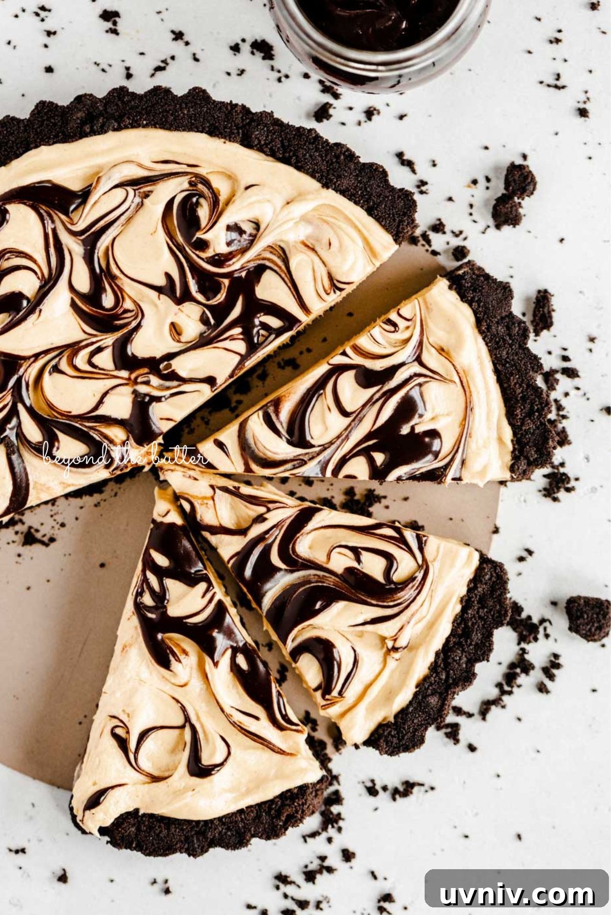 Round chocolate peanut butter tart swirled with chocolate syrup and cut into slices with a bowl of chocolate sandwich crumbs next to it on a white speckled background.
